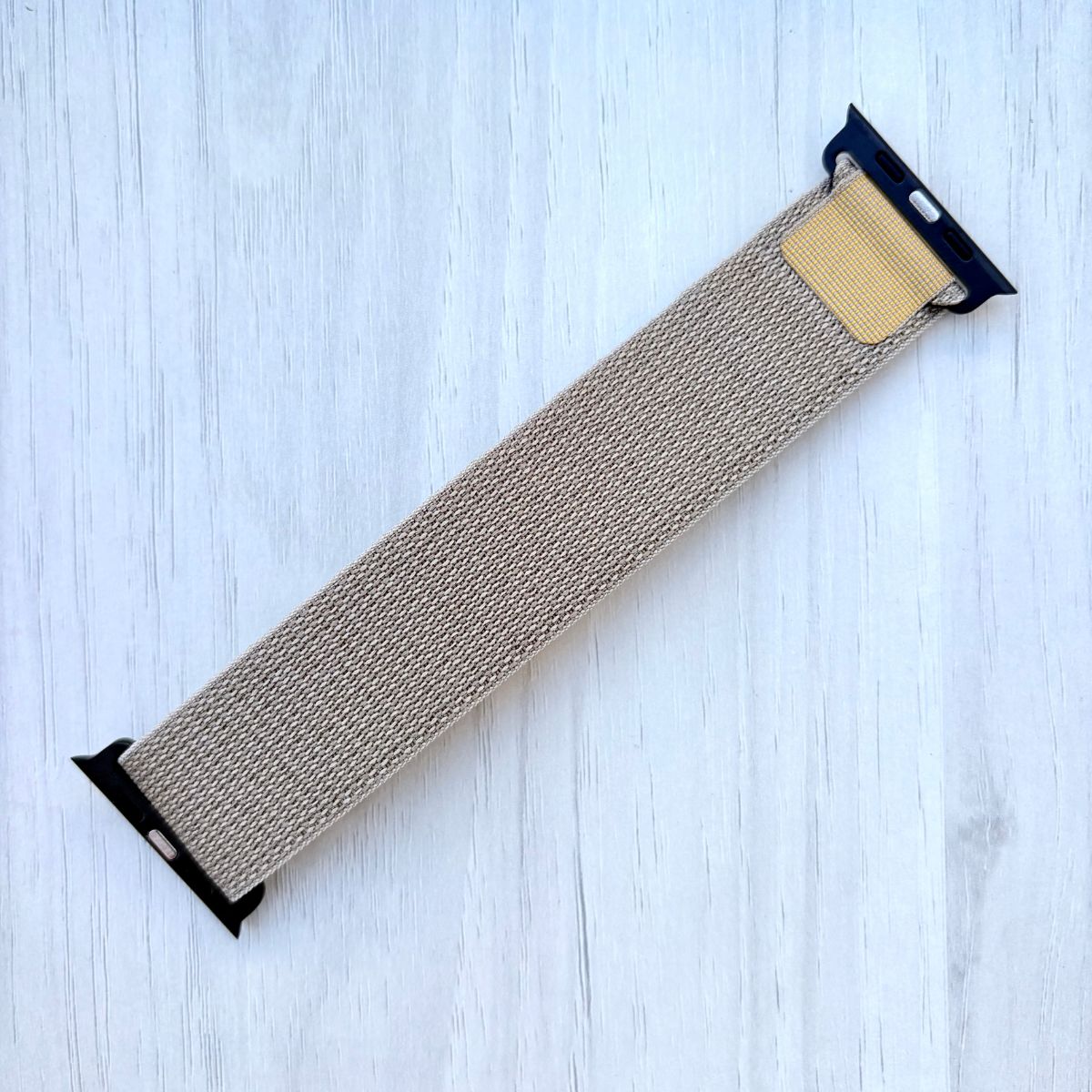 Tan Alpine Loop Apple Watch strap with black ends on a light blue wooden surface
