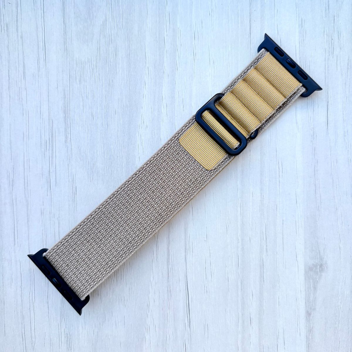 Tan Alpine loop Apple watch strap with black hardware on a light blue wooden background