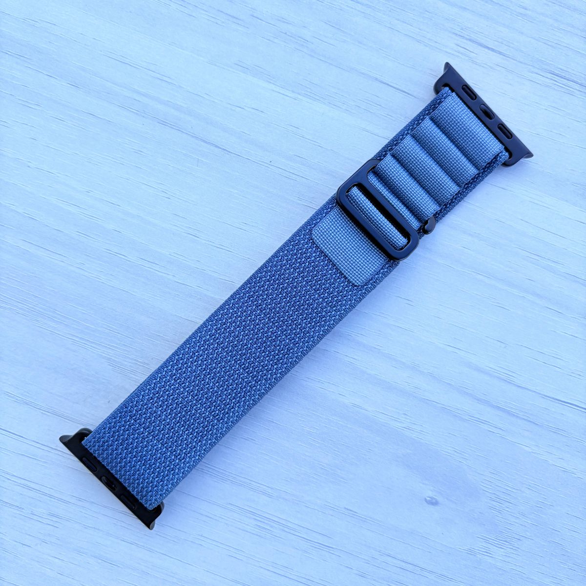 Light Blue Alpine Loop Apple Watch strap with black buckle on a light blue textured background
