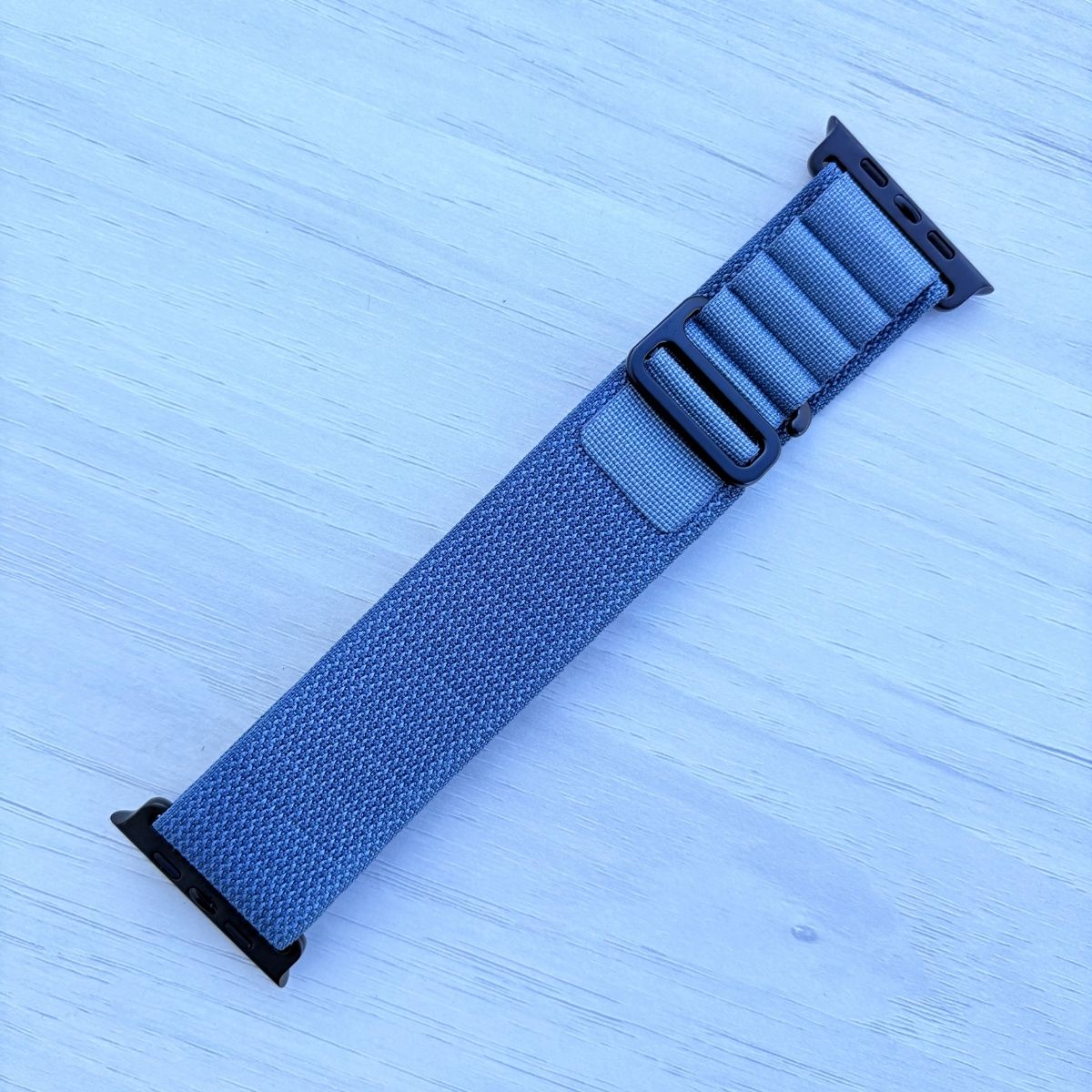 Light Blue Alpine loop Band for apple watch with Black finish on a white wooden background front view