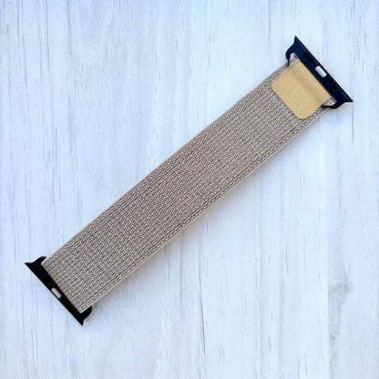 Tan Alpine Loop Apple Watch strap with black ends on a light blue wooden surface