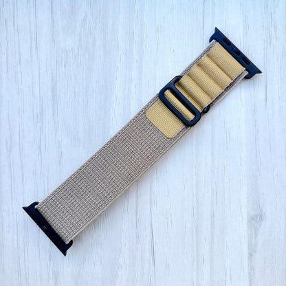 Tan Alpine loop Apple watch strap with black hardware on a light blue wooden background